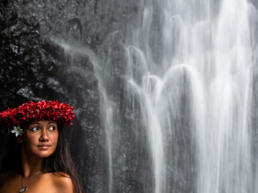 Frau mit Blumenkranz vor einem Wasserfall in tropischer Umgebung.