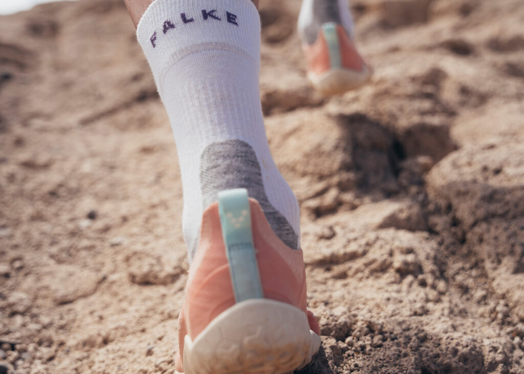 Nahaufnahme eines Trailrunning-Schuhs mit weißen Sportsocken auf sandigem Boden.