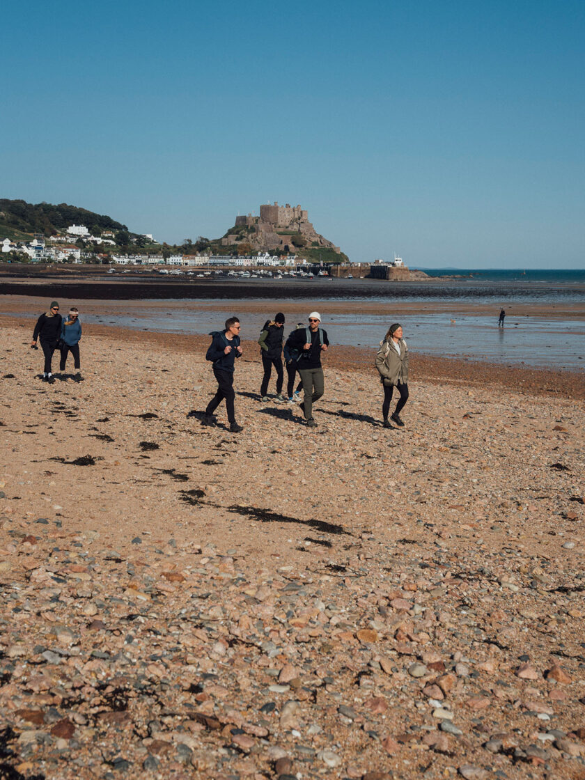 Menschen spazieren am Strand mit Blick auf eine Burg und blauen Himmel.