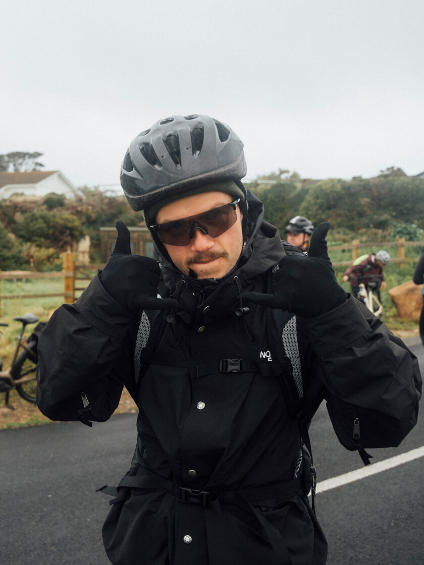 Radfahrer in schwarzer Kleidung und Helm zeigt Daumen-hoch-Geste auf einer Straße.