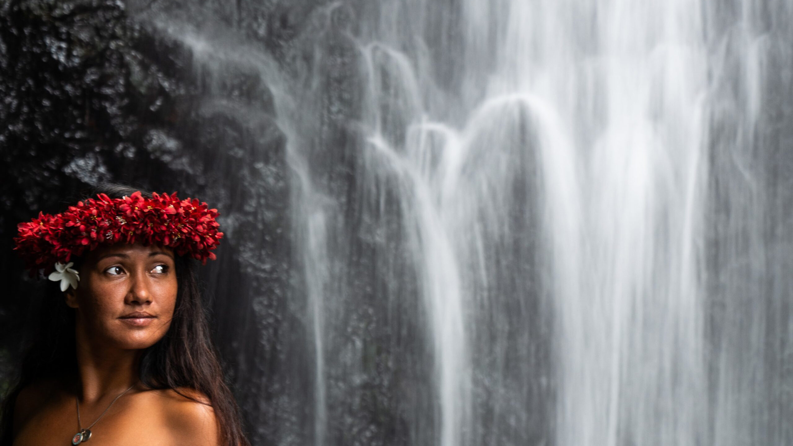 Frau mit Blumenkranz vor einem majestätischen Wasserfall in tropischer Umgebung.