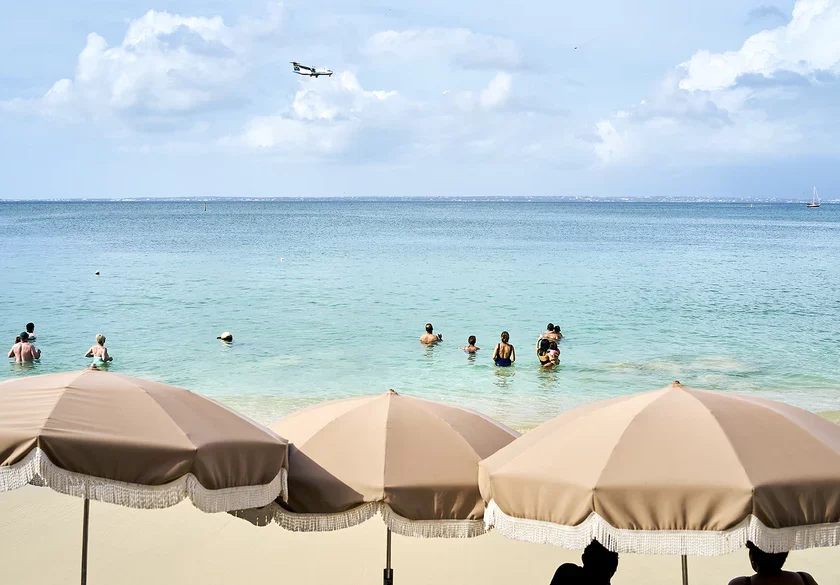 Strand mit Sonnenschirmen, Badegästen und einem Flugzeug am Himmel. Entspannende Urlaubsatmosphäre.