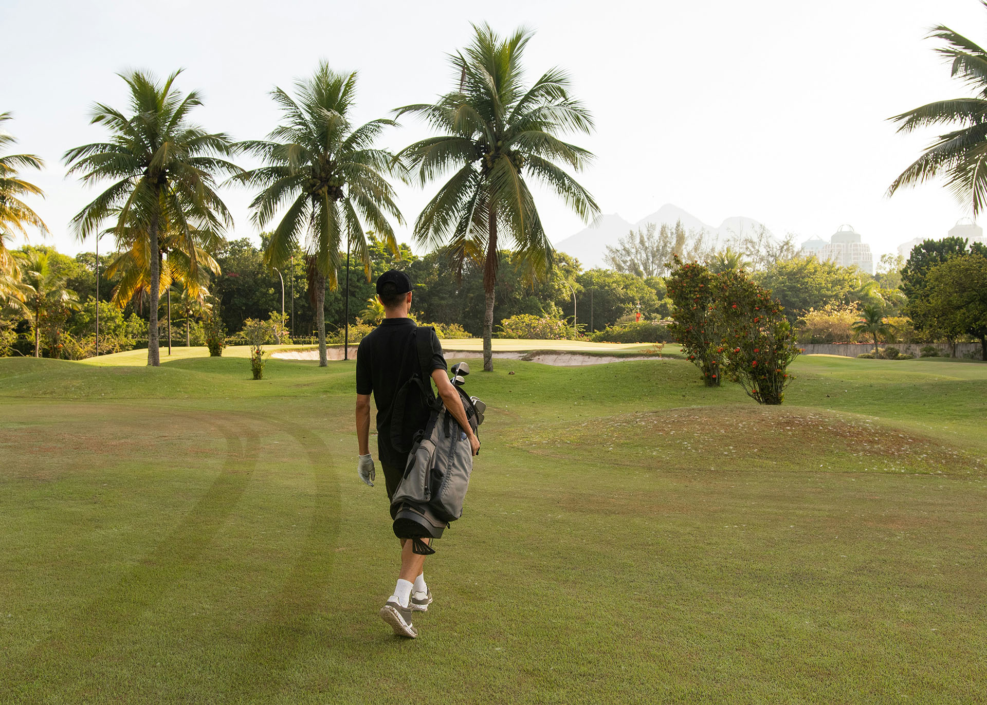 Golfspieler geht mit Golftasche über einen sonnigen Platz mit Palmen.