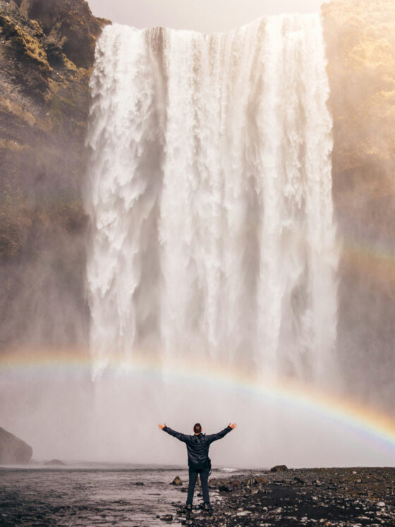 Person steht vor einem Wasserfall mit Regenbogen.