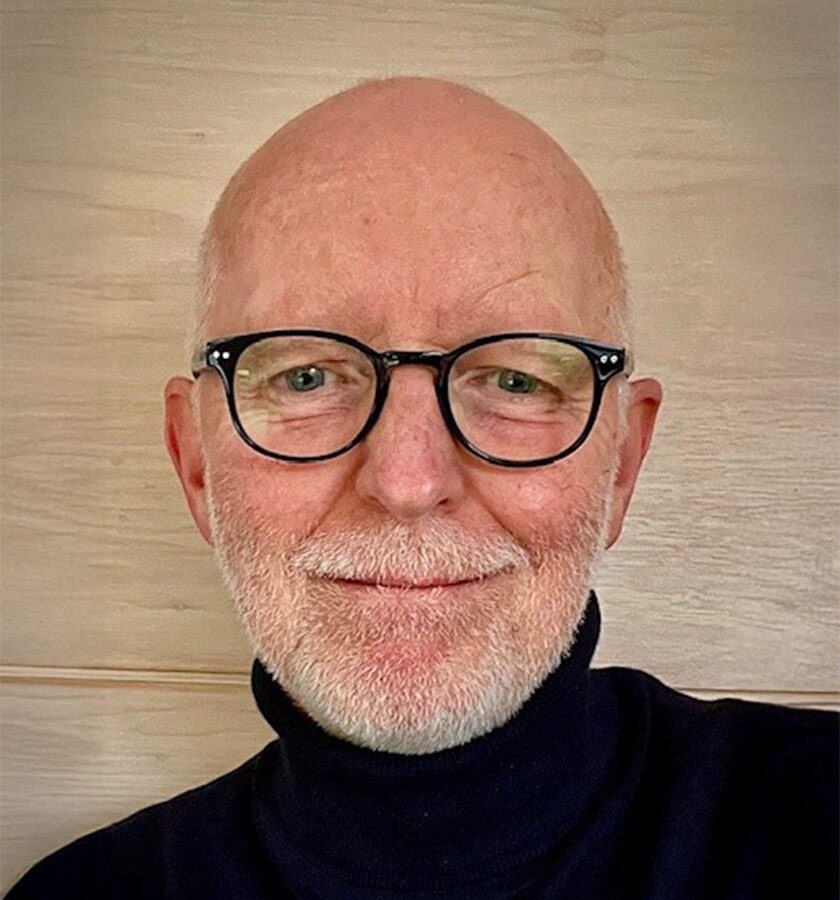 Porträt eines älteren Mannes mit Brille und Bart, lächelnd vor Holz-Hintergrund.